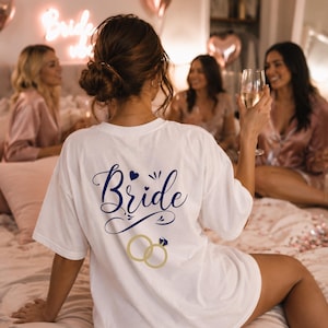 May include: A white t-shirt featuring the word Bride in dark blue cursive script, a small dark blue heart, and a graphic of two interlocking gold rings with a blue diamond. The design is printed on the back of the shirt, which is worn by a person.