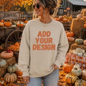 Może przedstawiać: Beżowa bluza z okrągłym dekoltem i napisem "ADD YOUR DESIGN" pogrubionymi pomarańczowymi literami. Odzież posiada długie rękawy, prążkowany dekolt oraz prążkowane mankiety. Bluza wykonana jest z jednolitego materiału o matowym wykończeniu.
