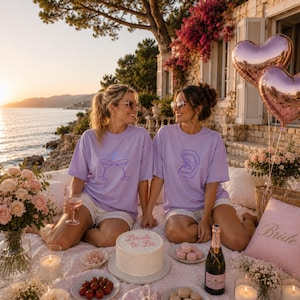 May include: Two people wearing matching light purple t-shirts, one with a graphic of two clinking martini glasses and the other with a ring box graphic. A white cake with the text Bride to Be sits on a blanket with flowers and champagne.
