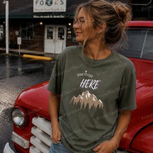 May include: Olive green t-shirt with the text "your design HERE" above a mountain and forest graphic. The person is wearing glasses and a necklace. A red vintage truck is in the background.