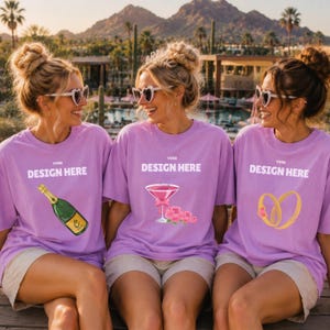 May include: Three light purple t-shirts featuring white text that reads YOUR DESIGN HERE. The left shirt displays a green champagne bottle, the center shirt shows a pink cocktail glass with flowers, and the right shirt features two gold rings.