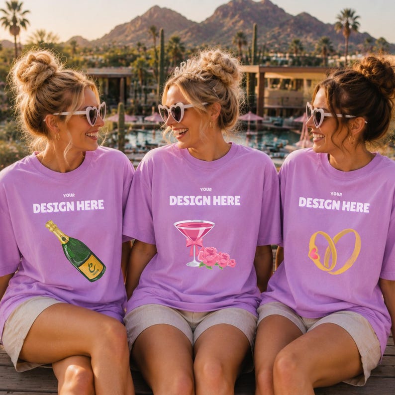May include: Three light purple t-shirts featuring white text that reads YOUR DESIGN HERE. The left shirt displays a green champagne bottle, the center shirt shows a pink cocktail glass with flowers, and the right shirt features two gold rings.
