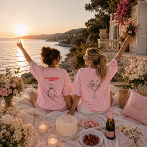 May include: Two light pink t-shirts featuring graphic prints on the back. One shirt displays a black line drawing of a diamond ring with the red text Your Design Here. The other shirt features a black line drawing of a person in a martini glass.