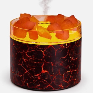 Puede incluir: Un difusor de aceites esenciales cilíndrico con un diseño de lava. La base es de color marrón oscuro con patrones de grietas rojas, coronada por un depósito de vidrio transparente lleno de piedras de color naranja. Una luz amarilla ilumina las piedras y el vapor se eleva desde el centro.
