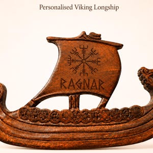 Op de afbeelding: Een donkerbruin houten snijwerk van een Viking-langschip. Het zeil is gegraveerd met het Vegvisir-symbool en de naam RAGNAR. Het schip heeft een gebogen romp met cirkelvormige schilddetails en een uitgesneden dierenkop aan de boeg.