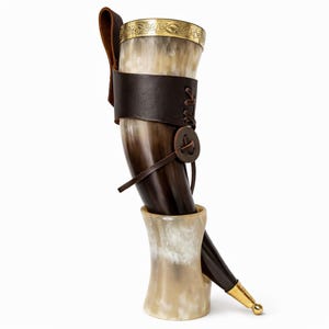 Könnte beinhalten: Ein Trinkhorn im Wikinger-Stil mit brauner Lederumwicklung und Riemen. Das Horn hat einen goldfarbenen Rand mit einem eingravierten Design. Es wird in einem passenden hornförmigen Ständer präsentiert. Das Horn ist etwa 30 cm hoch.