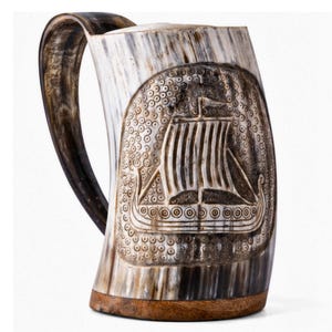 May include: A drinking mug crafted from a natural horn with a dark brown and cream-coloured finish. The exterior features a detailed carved relief of a sailing ship on a textured background. It includes a curved handle and a flat wooden base.