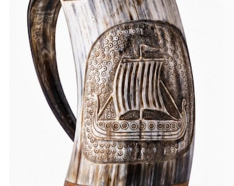 May include: A drinking mug crafted from a natural horn with a dark brown and cream-colored finish. The exterior features a detailed carved relief of a sailing ship on a textured background. It includes a curved handle and a flat wooden base.