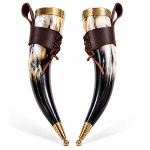 May include: Two curved drinking horns featuring polished black and white natural horn bodies. Each horn is fitted with a gold-toned metal rim with intricate engravings, a dark brown leather holder with lace-up detailing, and a gold-toned metal tip.