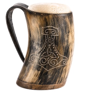 Könnte beinhalten: Ein geschwungener Trinkhornkrug mit dunkelbraun-beiger Marmorierung und einem eingravierten Mjolnir-Hammermotiv auf der Vorderseite. Der Krug hat einen dunklen Henkel und einen flachen Holzboden und ist mit einer dicken, hellen Schaumkrone gefüllt.