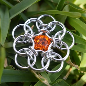 Könnte beinhalten: Eine silberne Kettenhemd-Blume mit einem orangefarbenen Edelstein in der Mitte.