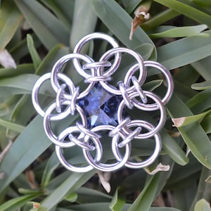May include: A silver metal flower charm with a blue gemstone in the center. The charm is made of interlocking rings.