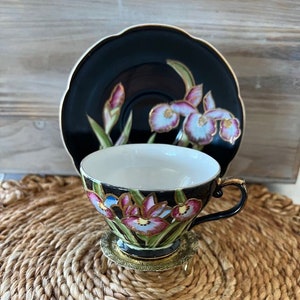 May include: A black and gold teacup and saucer set with a floral design featuring pink, blue, and green irises. The set is on a woven straw mat.