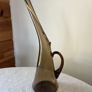 May include: A tall, brown glass pitcher with a curved spout and a handle. The pitcher has a unique, modern design.