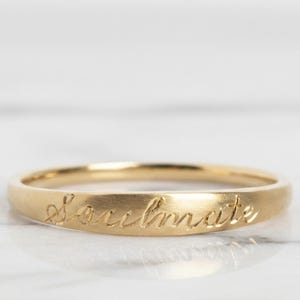 May include: A gold-colored ring with the word "Soulmate" engraved in a cursive font. The ring is thin and has a smooth, polished surface. The ring is set against a white marble background.