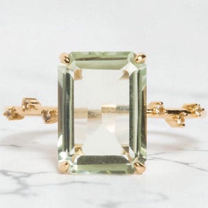 May include: A gold ring featuring a large, rectangular, light green gemstone. The ring has a delicate, branch-like design with small accent stones on either side of the main stone. The ring is set against a white marble background.