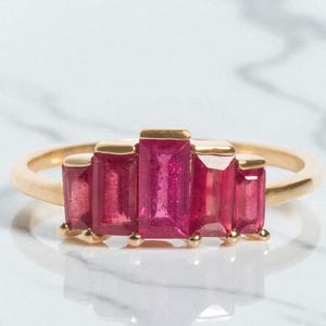 Könnte beinhalten: Ein goldener Ring mit fünf rechteckigen, leuchtend rosa Edelsteinen. Die Edelsteine sind in einer Reihe angeordnet, wobei der mittlere Stein etwas größer ist. Der Ring wird auf einer weißen Oberfläche präsentiert, die das Licht reflektiert.