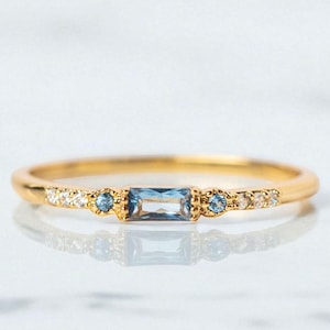 Pode incluir: Um delicado anel de ouro com uma pedra preciosa retangular azul clara como peça central. O anel é adornado com pedras azuis redondas menores e minúsculas pedras brancas. O anel é apresentado sobre um fundo branco, destacando seu design elegante.