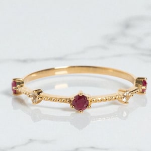 Puede incluir: Un delicado anillo de oro con pequeñas piedras preciosas redondas de color rojo oscuro y piedras claras más pequeñas. El anillo tiene una banda delgada con un diseño texturizado. Las piedras están espaciadas uniformemente.
