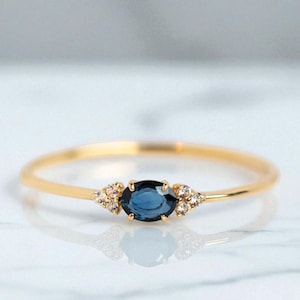 Può includere: Un delicato anello d'oro con una gemma ovale blu scuro al centro, affiancata da piccoli gruppi di pietre chiare. La fascia dell'anello è sottile e lucida. L'anello è un gioiello.