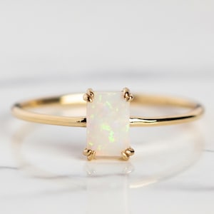 Puede incluir: Un anillo de oro con una piedra preciosa de ópalo rectangular. El ópalo tiene una base blanca con sutiles destellos verdes y amarillos. La banda del anillo es delgada y pulida, y la piedra está sujeta por cuatro garras.