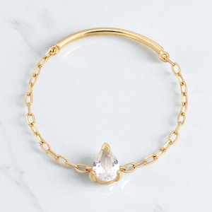 May include: A gold bracelet with a clear, pear-shaped gemstone at its centre. The bracelet has a chain-link design on the sides and a curved bar at the top. The gemstone is set in a gold bezel.