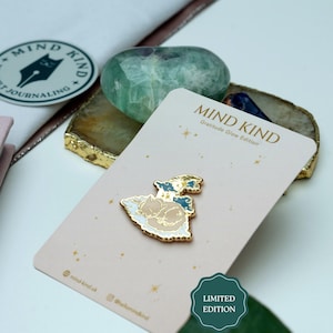 Può includere: Una spilla con un design di gatto addormentato in oro, bianco e blu, su una carta rosa con la scritta "MIND KIND Gratitude Glow Edition". Sono visibili anche una pietra verde e un adesivo con la scritta "MIND KIND".