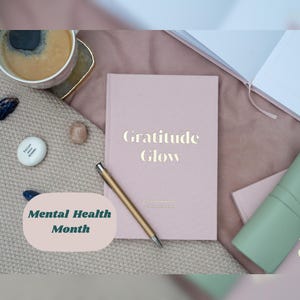 May include: A pink journal with gold lettering reading "Gratitude Glow", alongside a gold pen and a coffee cup. The image also features a small stone and a sign that says "Mental Health Month".