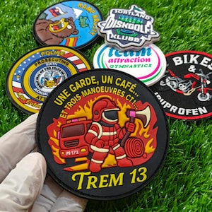 Op de afbeelding: Een verzameling geborduurde patches met diverse ontwerpen, waaronder een brandweerman met de tekst "TREM 13", een motorfiets en andere logo's. De patches zijn rond en kleurrijk, met een zwarte rand. Ze worden gepresenteerd op een groene ondergrond.