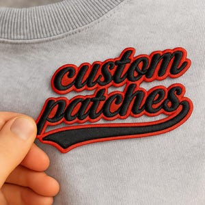 Peut inclure: Un sweat-shirt gris clair orné d'un écusson brodé portant le texte custom patches en lettres cursives noires, souligné par une bordure rouge vive. L'écusson présente un style athlétique classique avec un long trait sous le mot final.