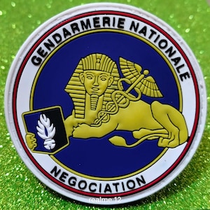 Peut inclure: Un écusson rond avec les mots "Gendarmerie Nationale" et "Négociation". Il présente une figure de sphinx dorée sur fond bleu, une bordure blanche et rouge, et un caducée. Le design est circulaire et détaillé.