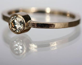 May include: A gold ring with a round, clear gemstone set in a bezel setting. The band is a simple, polished gold. The ring is displayed on a reflective surface, creating a mirrored effect. The ring is a piece of jewelry.