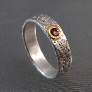 Puede incluir: Anillo de plata con una banda texturizada y un engaste de oro que sostiene una pequeña gema redonda y púrpura. El anillo tiene una textura martillada en la banda exterior y un interior liso y pulido.