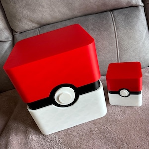 May include: Two novelty boxes in the style of a Pokeball, red and white with a black band and white central button. The larger box is angled, the smaller one upright. The boxes are likely for storage.
