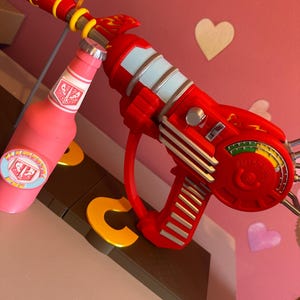 May include: A red toy ray gun with light blue detailing and a pink bottle with a label. The ray gun has a handle and dials. The bottle is pink with a white and red label.