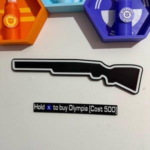 Puede incluir: Decoración de pared en forma de pistola en blanco y negro con borde blanco. Encima de la pistola hay un letrero que dice "Hold x to buy Olympia". La imagen también muestra tres estantes hexagonales con botellas, cada uno con un esquema de color diferente.