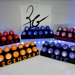 Peut inclure: Présentation de jetons de jeu de collection de différentes couleurs : rouge, orange, bleu, violet et rose. Chaque jeton présente un graphisme unique et est étiqueté avec des mots tels que "ULTRA" ou "EPIC". Les jetons sont disposés sur des présentoirs noirs à plusieurs niveaux.