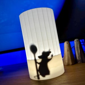 May include: A white, pleated cylindrical lamp, internally lit, projecting a shadow of a cartoon figure with a spoon. Three small decorative figures are positioned to the right of the lamp.