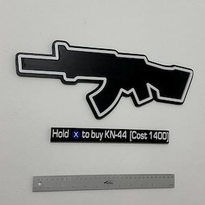 Può includere: Un'insegna da parete in bianco e nero a forma di sagoma di fucile, accompagnata da una targa rettangolare con la scritta "Hold X to buy KN-44 [Cost 1400]" e un righello metallico da 30 cm posizionato sotto per mostrare la scala.