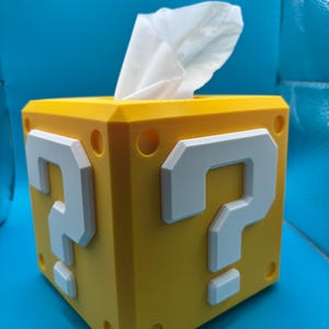 May include: A yellow tissue box featuring white question marks and a white tissue protruding from the top. The cube-shaped box has rounded corners and small circular details. The design is inspired by a video game.