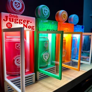 Op de afbeelding: Een display van vier frisdrankautomaten met verlichte borden en blikjes. De eerste machine is rood met de tekst "JUGGER-NOG" en een rood blikje. De tweede is groen met de tekst "Speed Cola" en een groen blikje. De derde is oranje met de tekst "Double Tap Root Beer" en een oranje blikje. De vierde is blauw.
