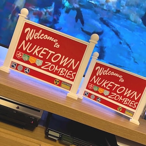 Pode incluir: Duas placas com moldura branca com fundos vermelhos e o texto "Welcome to Nuketown Zombies". As placas têm elementos decorativos e são colocadas sobre uma superfície de madeira. As placas estão em frente a um fundo azul.