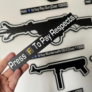 May include: A black sticker with the text "Press F To Pay Respects" in white. The letter "F" is in a yellow square. Silhouettes of black guns are in the background, with text saying "Hold x to buy Ray Gun" and "Hold x to buy MP40".