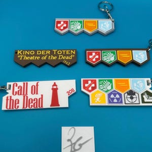 May include: A collection of keychains with designs from the game Call of Duty: Zombies. The keychains feature text such as "KINO DER TOTEN" and "Call of the Dead", along with various graphic elements. The keychains are white, brown, and multi-colored.