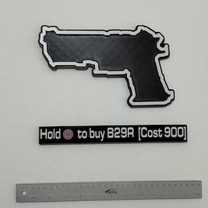 May include: A black 3D-printed handgun silhouette with a white border, accompanied by a black rectangular sign with white text reading "Hold [square icon] to buy B29R [Cost 900]" and a 30 cm metal ruler for scale, showing the gun is about 20 cm long.
