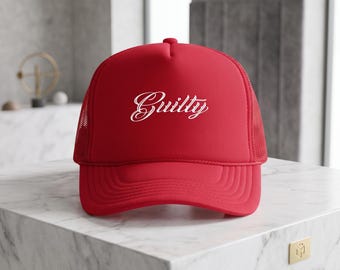 May include: A bright red trucker hat featuring a mesh back and a curved brim. The front panel displays the word Guilty in a white, cursive script font. The hat is constructed with a structured foam front and a matching red plastic snapback closure.