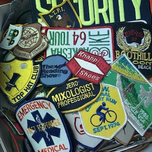 May include: A collection of embroidered patches with various designs and text. Patches include "SECURITY", "EMERGENCY FIRST RESPONDER", "Knapp Shoes", and "Bicycle USA 1986". Colors include blue, yellow, red, and green.