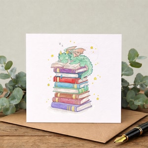 Peut inclure: Carte de vœux carrée avec une illustration à l'aquarelle d'un dragon vert dormant sur une pile de livres colorés. Le dragon a des cornes et des ailes jaunes. Des étoiles jaunes sont dispersées autour du dragon.