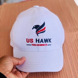 May include: White baseball cap with the text "US HAWK" in red and black, and a stylized bird logo. The cap also features the phrase "YOU DESERVE IT" in black.