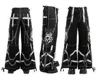 May include: Black cargo pants with white stitching, straps, and chains. The pants feature multiple pockets and decorative tribal designs. The wide-leg design is accented with white trim and chains. The pants are displayed from multiple angles.
